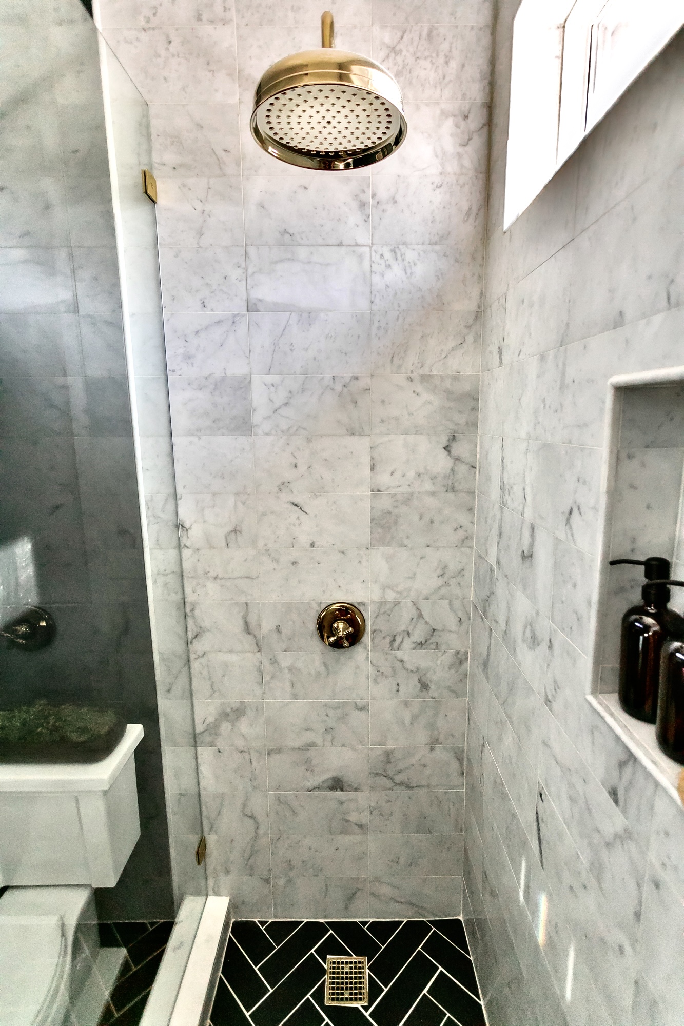Marble walk-in shower with brass rain head, built-in niche, and black herringbone tile floor — J. Arthur Custom Homes